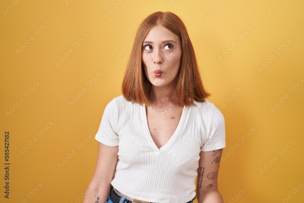 Poster young redhead woman standing over yellow background making fish face with lips, crazy and comical ge