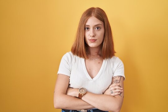 Young redhead woman standing over yellow background skeptic and nervous, disapproving expression on face with crossed arms. negative person.