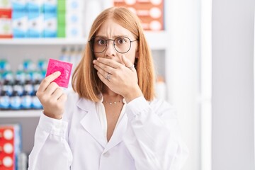 Young redhead woman working at pharmacy drugstore holding condom covering mouth with hand, shocked...