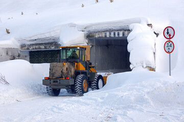 A large tractor clears snow from the entrance to a narrow tunnel. Snowbound narrow country road going through rectangular concrete tunnel and great snowdrifts.