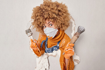 Photo of surprised curly haired female construction worker holds paint brushes improves house or apartment wears protective respirator jumper gloves poses in hole of plaster wall. Occupation concept