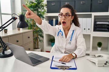 Young hispanic woman wearing doctor uniform and stethoscope pointing with finger surprised ahead, open mouth amazed expression, something on the front