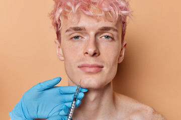 Horizontal shot of serious young pink haired guy receives injection in chin poses in beauty clinic...