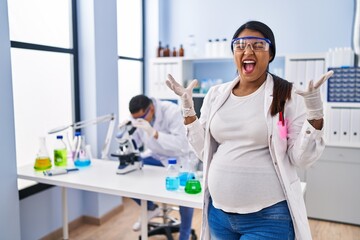 Young hispanic woman expecting a baby working at scientist laboratory celebrating mad and crazy for success with arms raised and closed eyes screaming excited. winner concept