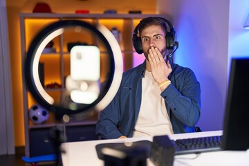 Hispanic man with beard recording vlog tutorial with smartphone at home covering mouth with hand,...