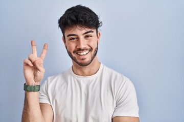 Hispanic man with beard standing over white background smiling with happy face winking at the...