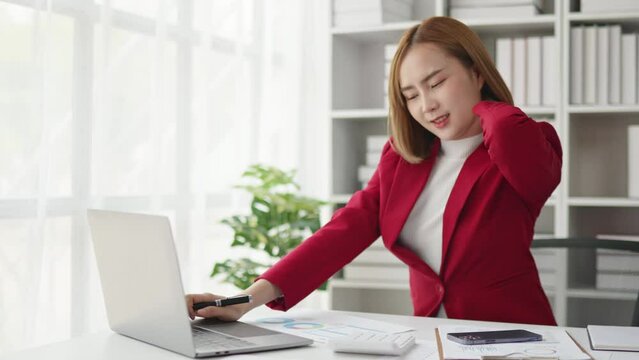 Business Woman in red suit seriously stressed working and neck pain and shoulder pain due to office syndrome on laptop and paperwork in the office.