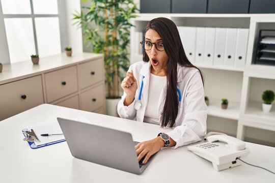 Young hispanic doctor woman doing video call at the clinic scared and amazed with open mouth for surprise, disbelief face