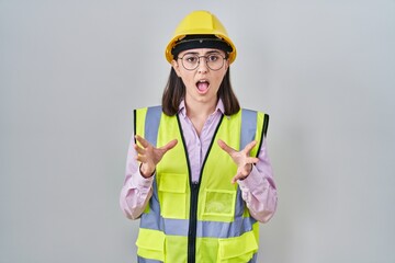 Hispanic girl wearing builder uniform and hardhat crazy and mad shouting and yelling with aggressive expression and arms raised. frustration concept.