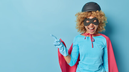 Overjoyed curly female model dressed in superhero costume with helmet eyemask and cloak points...