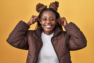 African woman with braided hair standing over yellow background smiling pulling ears with fingers,...