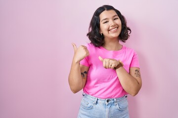 Young hispanic woman standing over pink background pointing to the back behind with hand and thumbs up, smiling confident