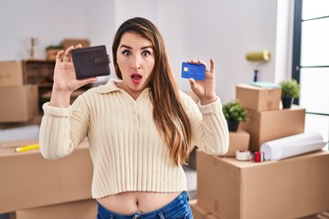 Young hispanic woman moving to a new home holding wallet and credit card in shock face, looking...