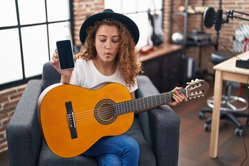 Young caucasian woman playing classic guitar at music studio holding smartphone making fish face with mouth and squinting eyes, crazy and comical.