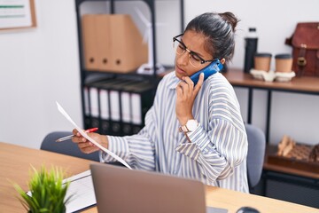 Young beautiful hispanic woman business worker talking on smartphone writing on document at office