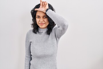 Hispanic woman with dark hair standing over isolated background making fun of people with fingers on forehead doing loser gesture mocking and insulting.