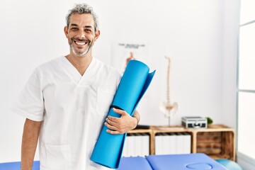 Middle age grey-haired man physiotherapist smiling confident holding yoga mat at rehab clinic