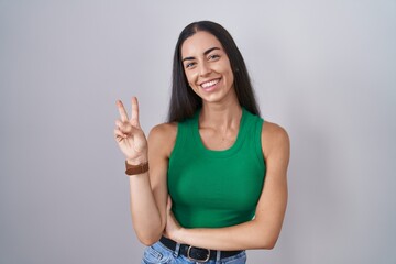 Young woman standing over isolated background smiling with happy face winking at the camera doing victory sign with fingers. number two.