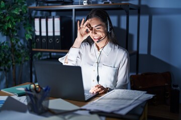 Young brunette woman wearing call center agent headset working late at night doing ok gesture with hand smiling, eye looking through fingers with happy face.