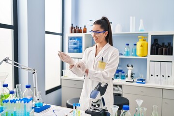 Young beautiful hispanic woman scientist smiling confident wearing gloves at laboratory