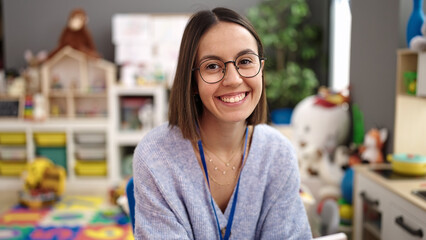 Young beautiful hispanic woman teacher smiling confident sitting on table at kindergarten