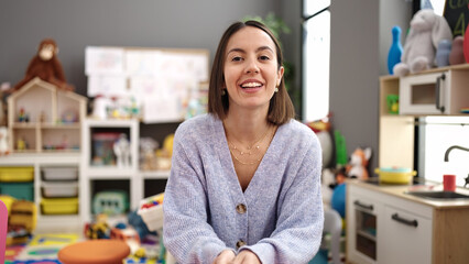 Young beautiful hispanic woman teacher smiling confident sitting on table at kindergarten