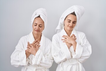 Middle age woman and daughter wearing white bathrobe and towel smiling with hands on chest with closed eyes and grateful gesture on face. health concept.