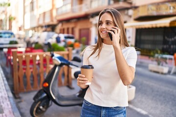 Young blonde woman talking on the smartphone and drinking coffee at street