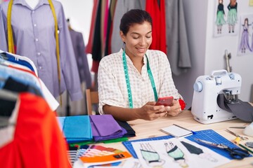 Young beautiful hispanic woman tailor smiling confident using smartphone at tailor shop