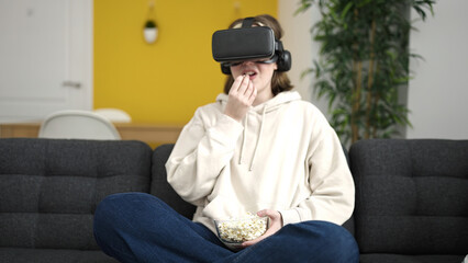 Fototapeta premium Young blonde woman watching movie using virtual reality glasses eating popcorn at home