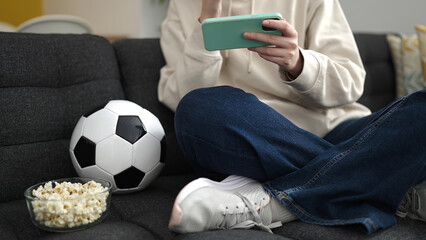 Young blonde woman watching soccer match on smartphone sitting on sofa at home