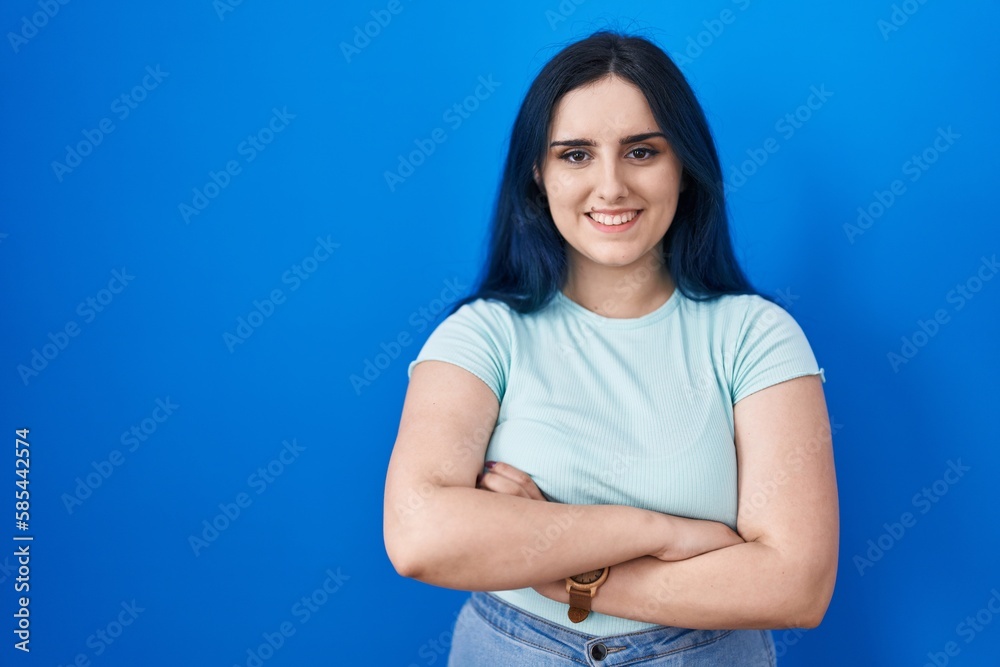 Wall mural Young modern girl with blue hair standing over blue background happy face smiling with crossed arms looking at the camera. positive person.