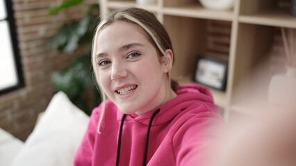 Young blonde woman make selfie by camera sitting on bed at bedroom