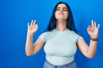Fototapeta premium Young modern girl with blue hair standing over blue background relaxed and smiling with eyes closed doing meditation gesture with fingers. yoga concept.