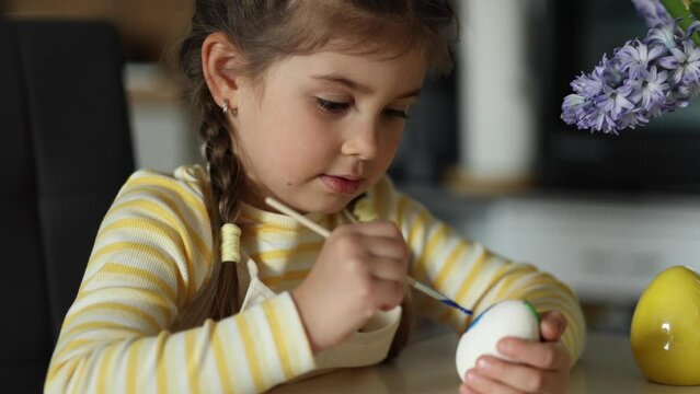Cute four year girl sitting on kitchen table and painting easter eggs. Yellow and blue colour. Ukrainian symbolic 