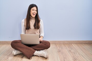 Young brunette woman working using computer laptop sitting on the floor winking looking at the...