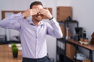 Young hispanic man at the office covering eyes with hands smiling cheerful and funny. blind concept.