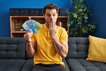 Young hispanic man holding chilean pesos covering mouth with hand, shocked and afraid for mistake....