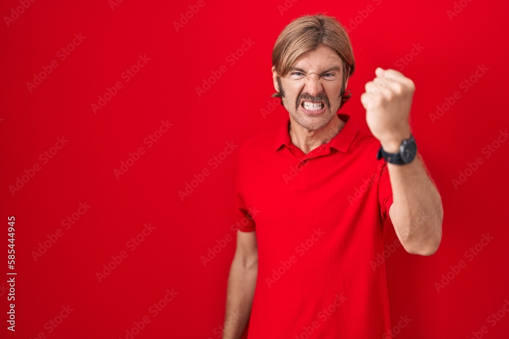 Wall mural Caucasian man with mustache standing over red background angry and mad raising fist frustrated and furious while shouting with anger. rage and aggressive concept.