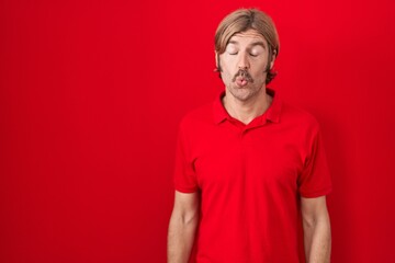 Fototapeta premium Caucasian man with mustache standing over red background making fish face with lips, crazy and comical gesture. funny expression.