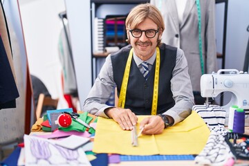 Young blond man tailor smiling confident make mark on cloth at clothing factory