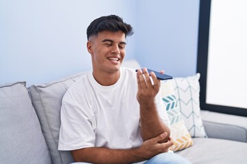 Young hispanic man talking on the smartphone sitting on sofa at home