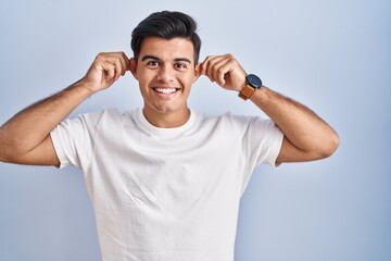 Fototapeta premium Hispanic man standing over blue background smiling pulling ears with fingers, funny gesture. audition problem