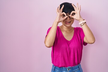 Young asian woman with short hair standing over pink background doing ok gesture like binoculars sticking tongue out, eyes looking through fingers. crazy expression.