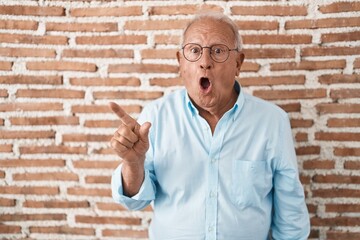 Senior man with grey hair standing over bricks wall surprised pointing with finger to the side, open mouth amazed expression.