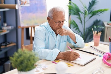 Senior grey-haired man artist drawing on notebook with doubt expression at art studio