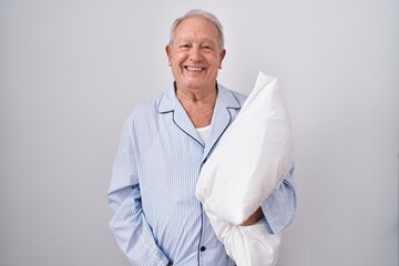 Senior man with grey hair wearing pijama hugging pillow with a happy and cool smile on face. lucky person.