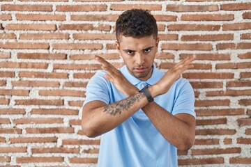 Brazilian young man standing over brick wall rejection expression crossing arms doing negative sign, angry face