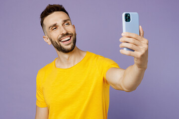 Young smiling caucasian man wear yellow t-shirt doing selfie shot on mobile cell phone post photo on social network isolated on plain pastel light purple background studio portrait. Lifestyle concept.