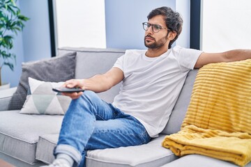 Handsome latin man holding television remote control relaxed with serious expression on face....